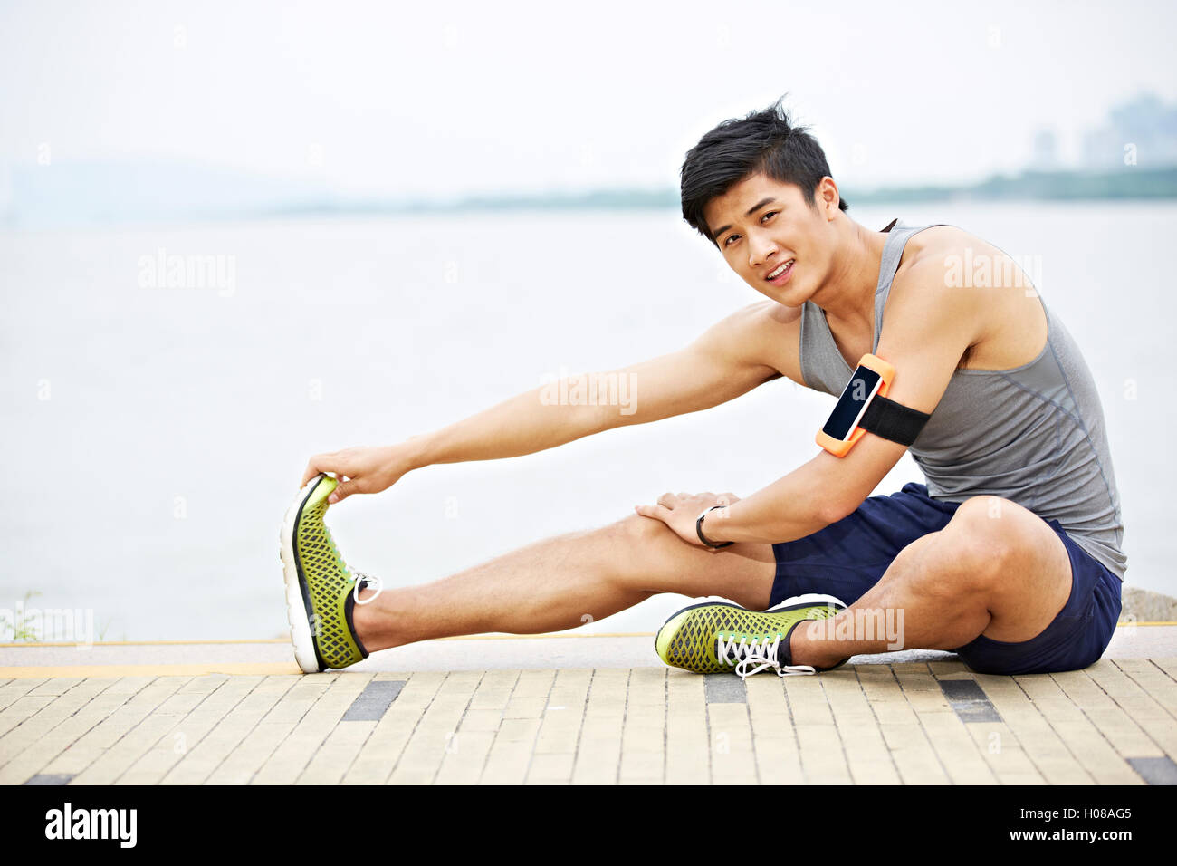 young handsome asian jogger wearing fitness tracker stretching a leg by lake looking at camera smiling Stock Photo