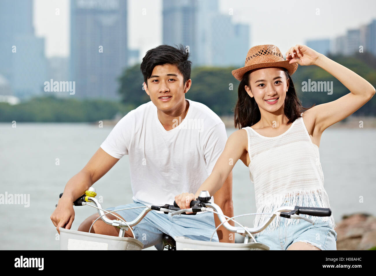 young asian couple riding bike in urban park, looking at camera smiling Stock Photo