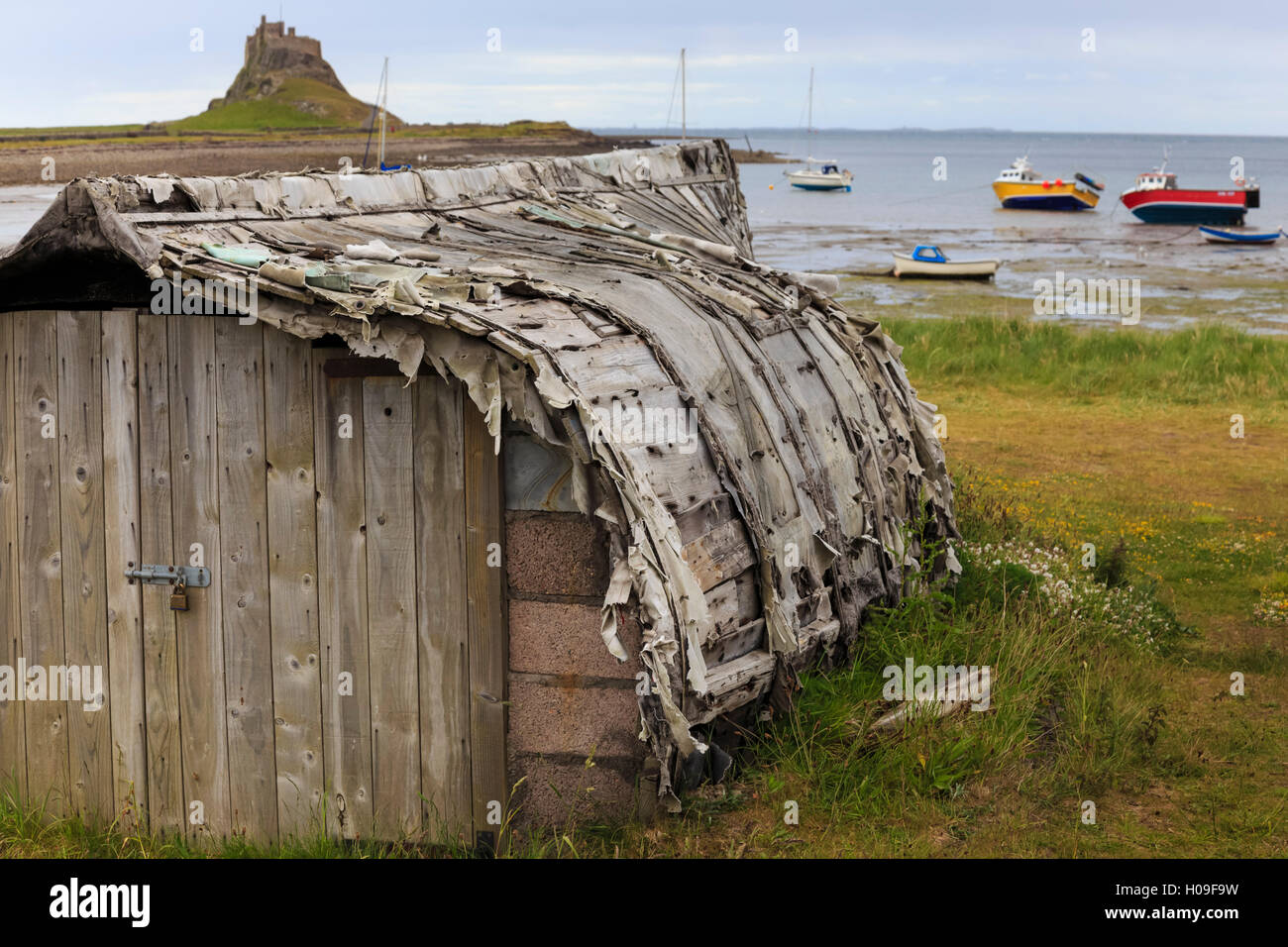 Upturned weathered boat hut with Lindisfarne Castle and fishing boats at low tide, Holy Island, Northumberland Coast, UK Stock Photo