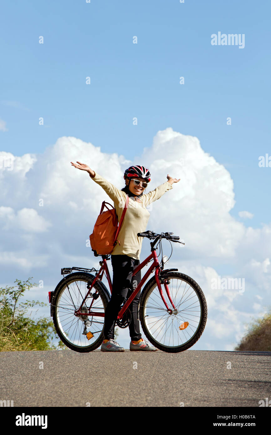 Woman cyclist standing with a bicycle on the road horizon and hands lifted up to heaven. Stock Photo