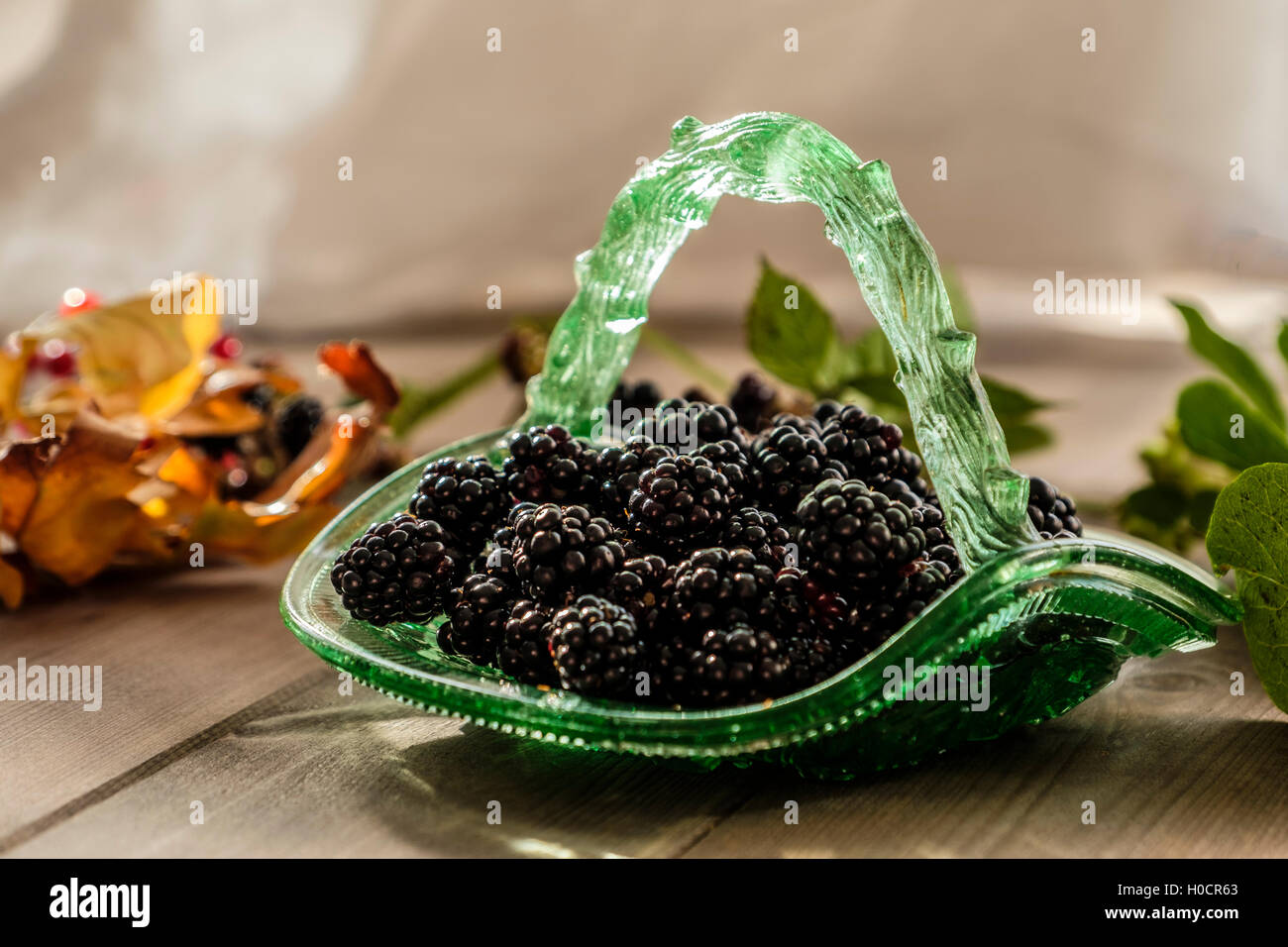 Vintage green glass basket full of blackberries with out of focus autumnal leaves on wooden table Stock Photo
