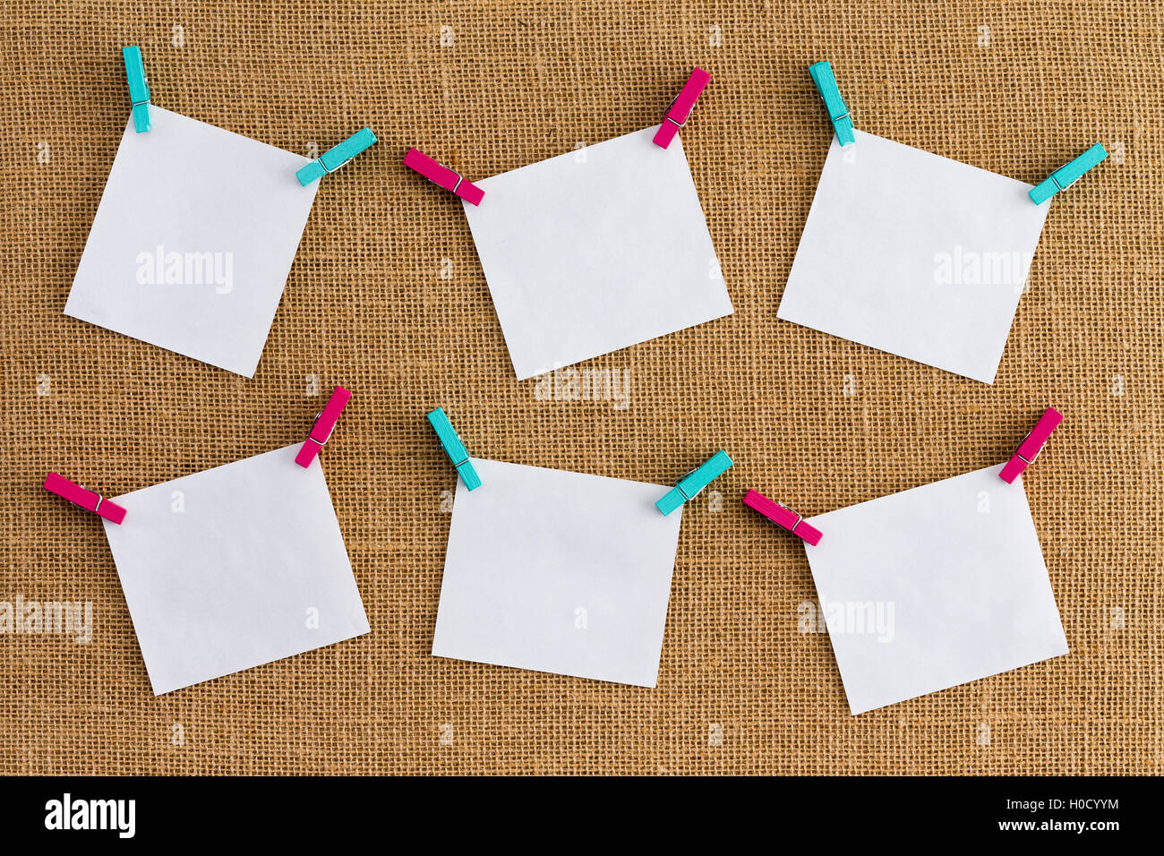 Six misaligned blank white notepads on hessian or burlap fabric hanging from alternating colorful pink and blue clothespins in a Stock Photo