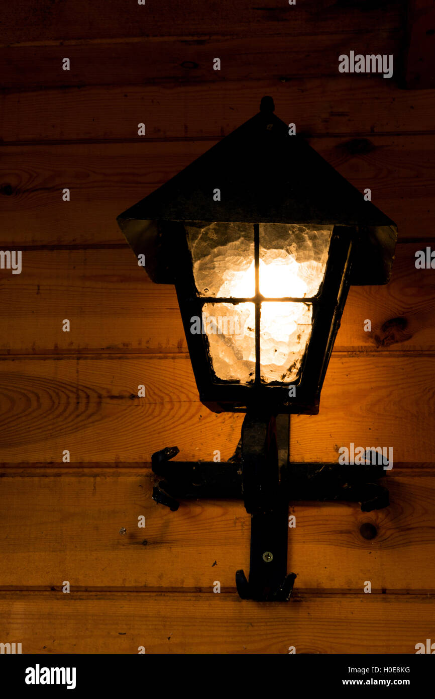 Shining, old metal lamp mounted on a wooden wall wooden building, summer evening. Vertical view. Stock Photo