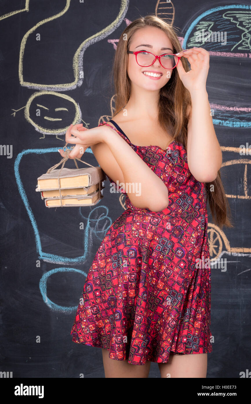 Young student girl in red dress with red glasses and pack of books poses in front of school classroom blackboard Stock Photo