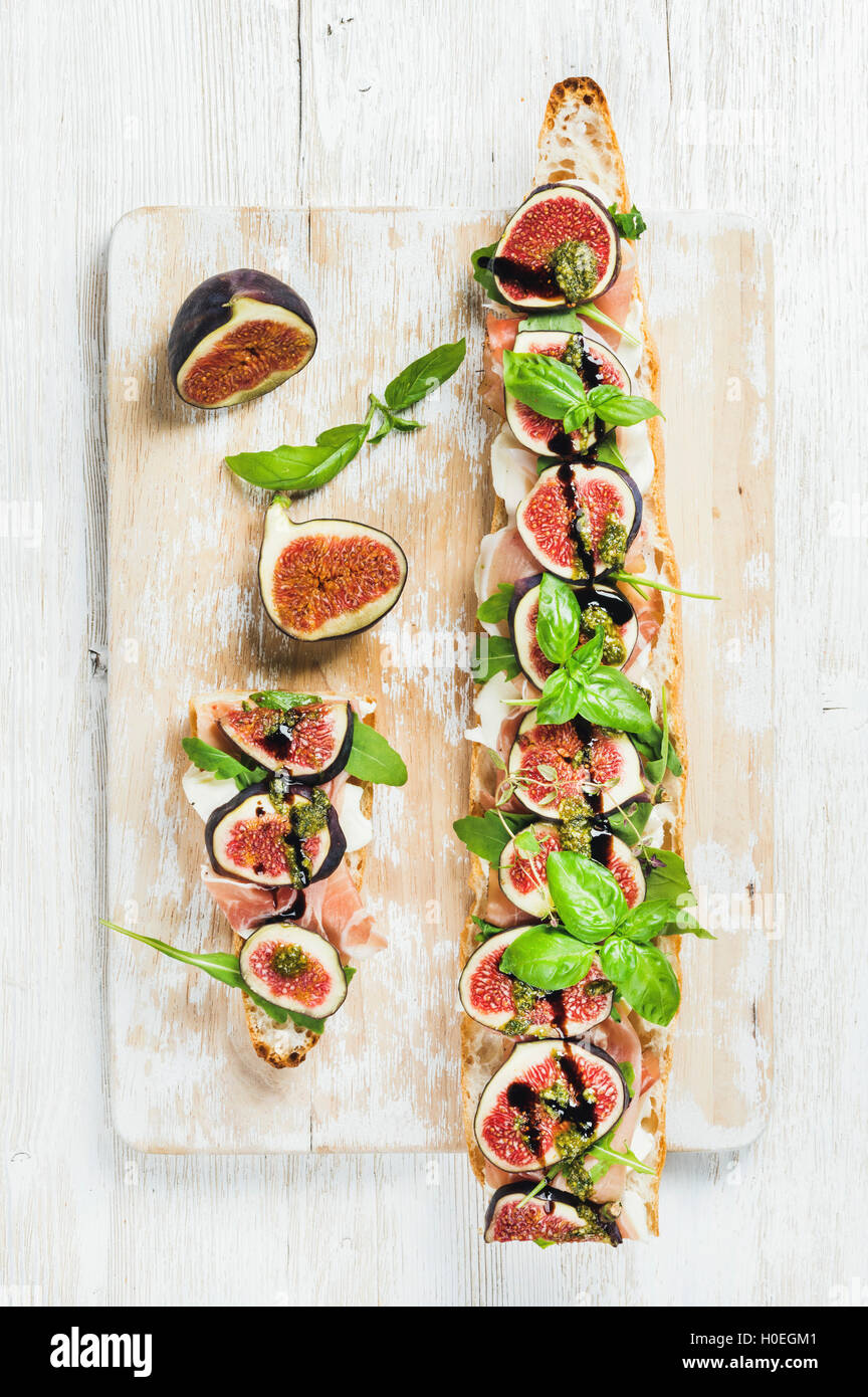 Long baguette sandwich with prosciutto meat, mozzarella cheese, arugula, figs and basil on shabby white painted wooden board ove Stock Photo