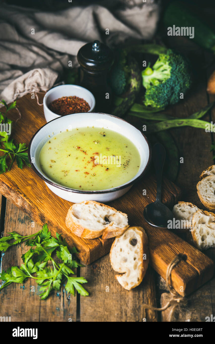 Fresh homemade pea, broccoli, zucchini cream soup in white bowl with fresh baguette slices and parsley on wooden board over rust Stock Photo