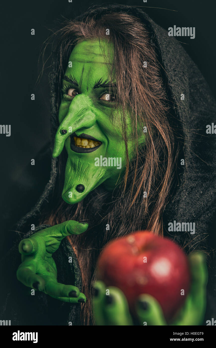 Green witch with long hair and dirty teeth holding a red apple in her hand. Shallow depth of field. Stock Photo
