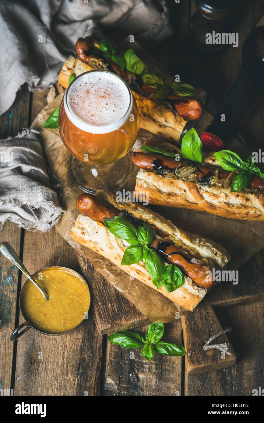Glass and bottle of wheat unfiltered beer and grilled sausage dogs in baguette with mustard, caramelised onion and herbs on serv Stock Photo