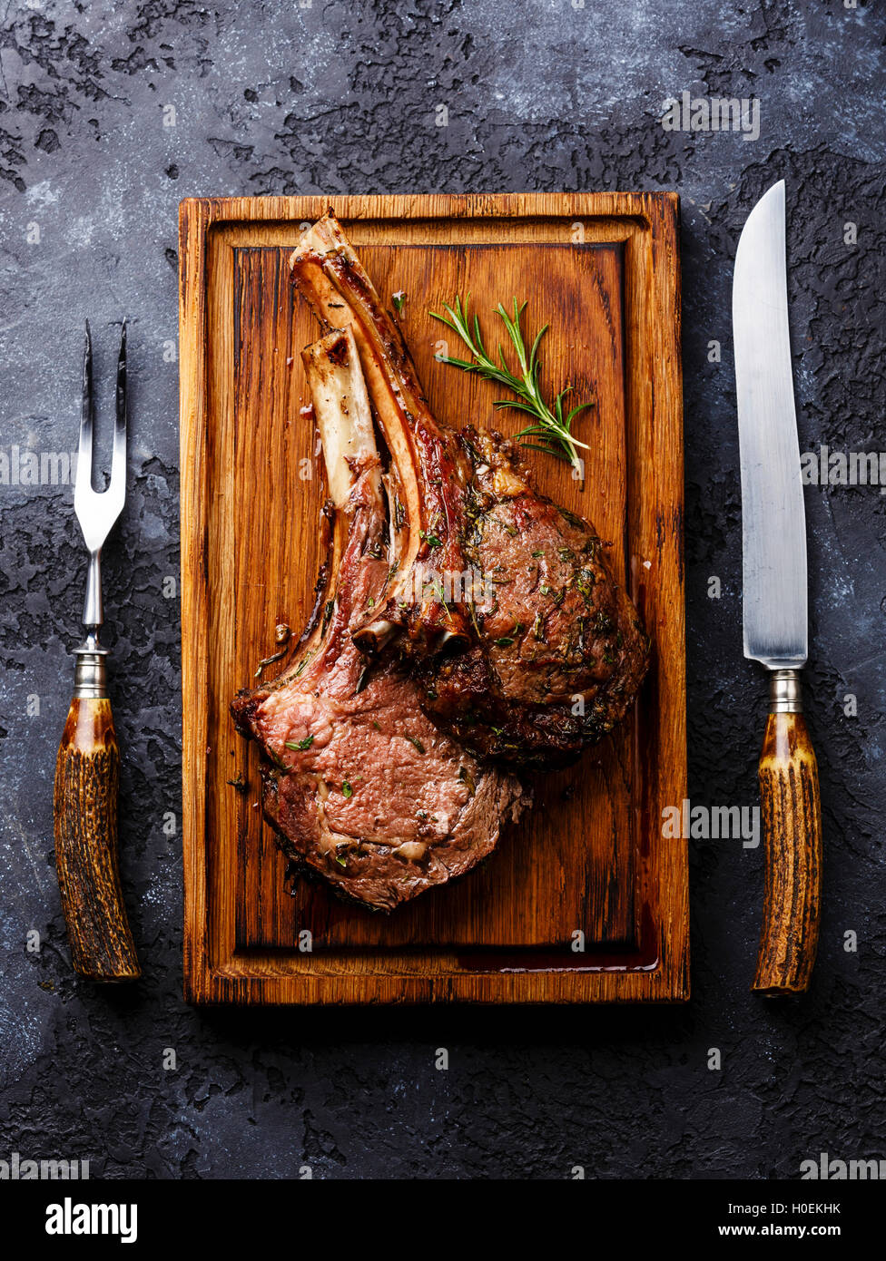 Roasted beef ribs on bone on wooden cutting board with knife and fork carving set on dark background Stock Photo
