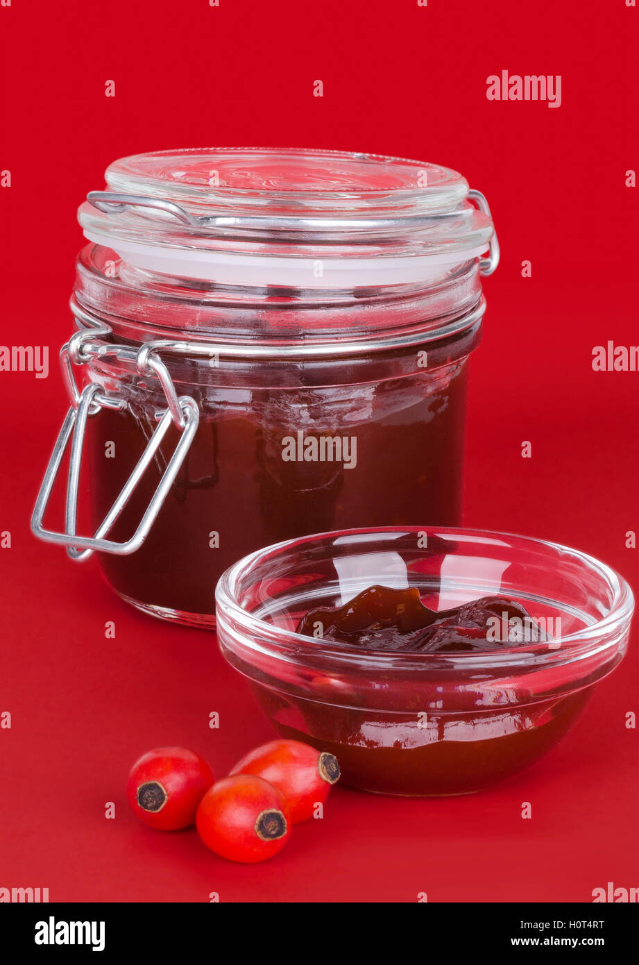 Rose hip jam and fruits over red background, also rose haw or rose hep. Marmelade in glass jar and bowl. Stock Photo