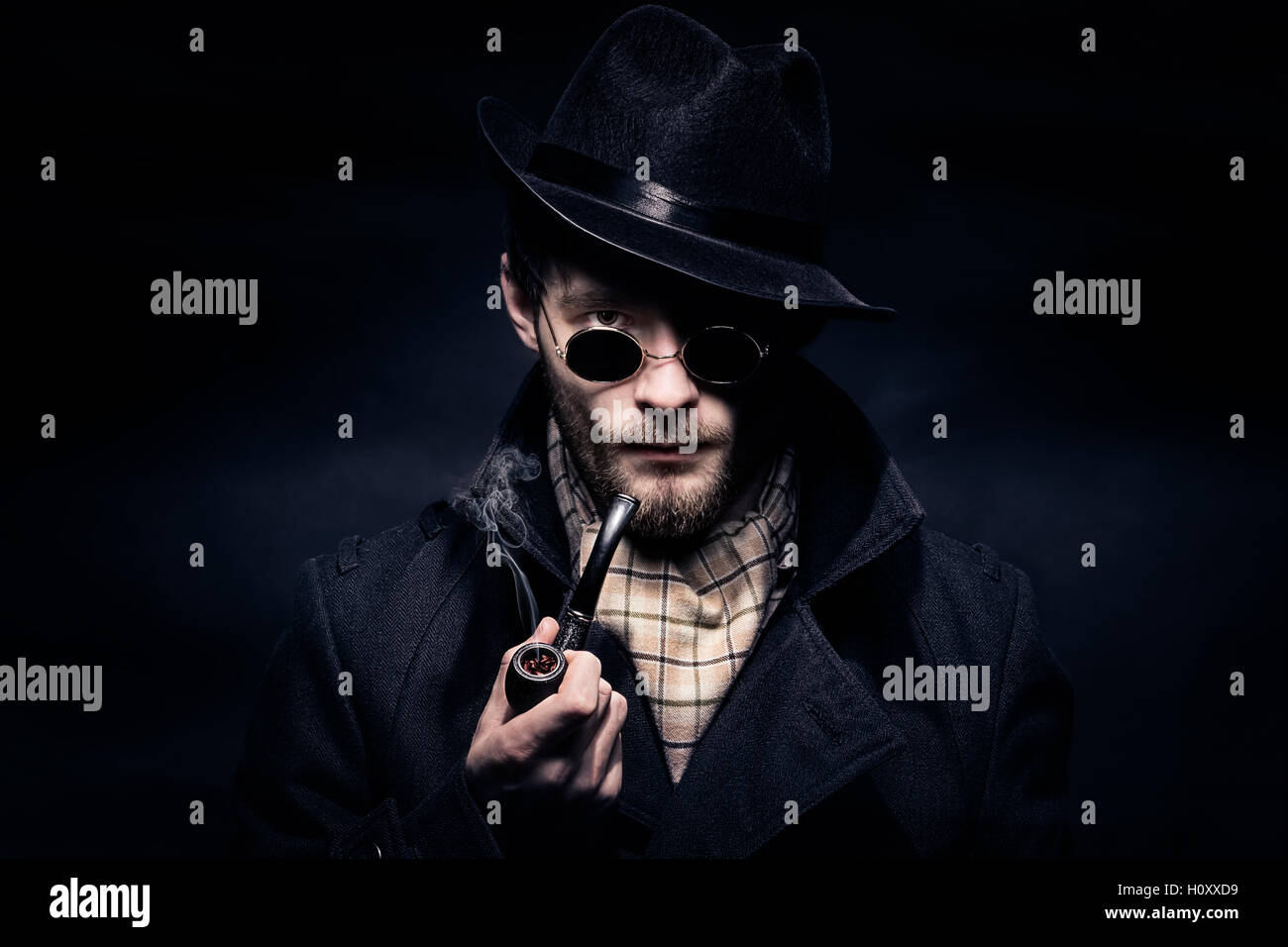 Portrait of a man, Sherlock Holmes like character on the black background. Holding a smoking pipe in the hand and wearing sunglasses, hat, scarf, coat Stock Photo