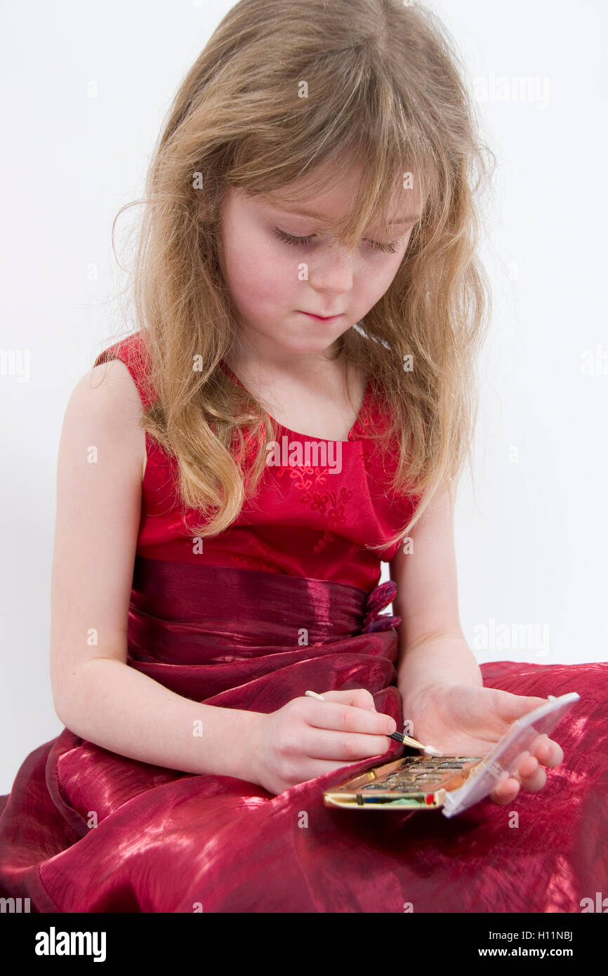 Playing with make-up : 6 year old girl wearing shiny red evening dress sitting  practicing applying eye shadow Stock Photo