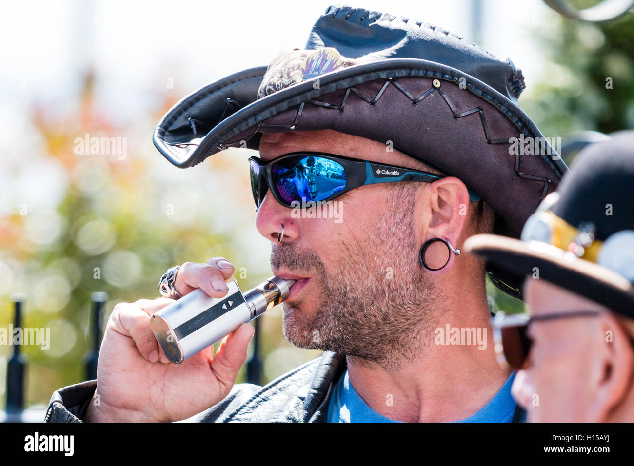 Caucasian man, unshaven and wearing cowboy hat and sunglasses, taking a puff from an e-cigarette, vape pen. Head and shoulder shot, side view. Stock Photo