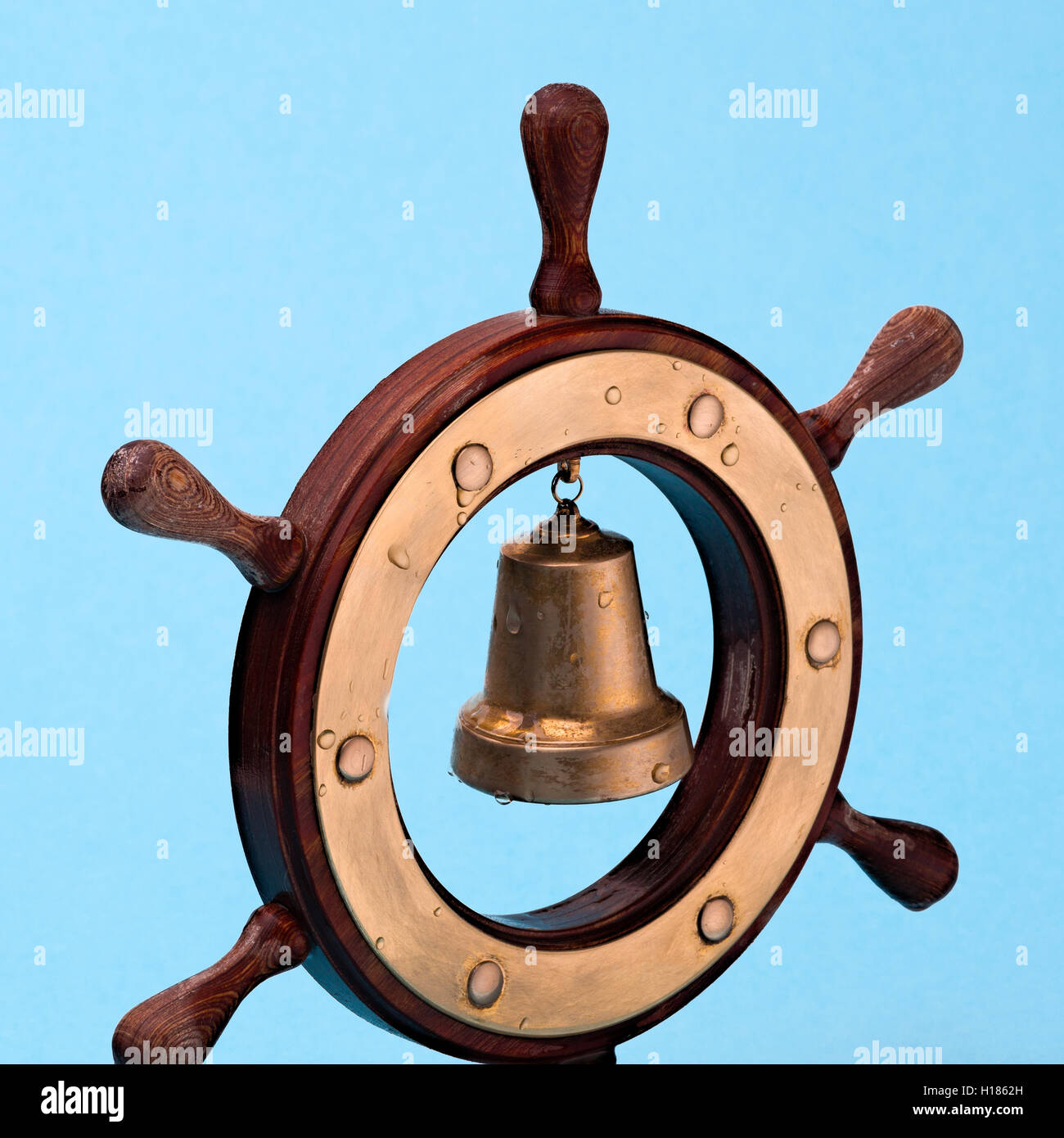 Decorative wood and brass toy helm or steering wheel and a ship's bell against pale blue background. Drops of sea water. Stock Photo