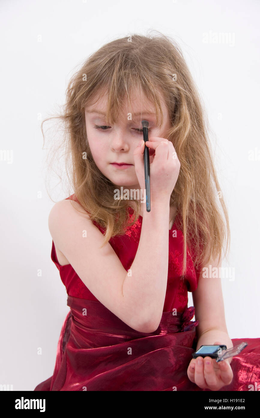 Playing with make-up : 6 year old girl wearing shiny red evening dress sitting  practicing applying eye shadow Stock Photo