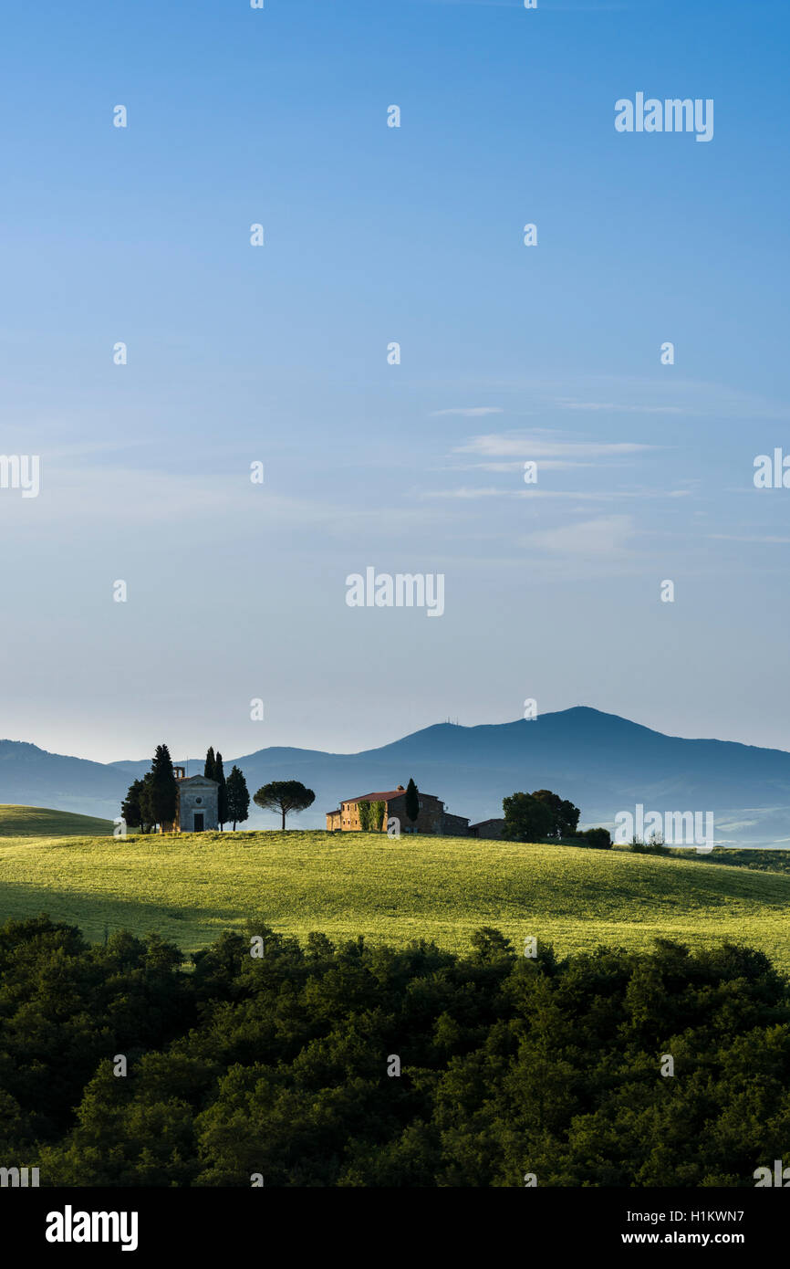Typical green Tuscan landscape in Val d’Orcia with farm and chapel on hill, fields, cypresses, trees and blue, cloudy sky Stock Photo