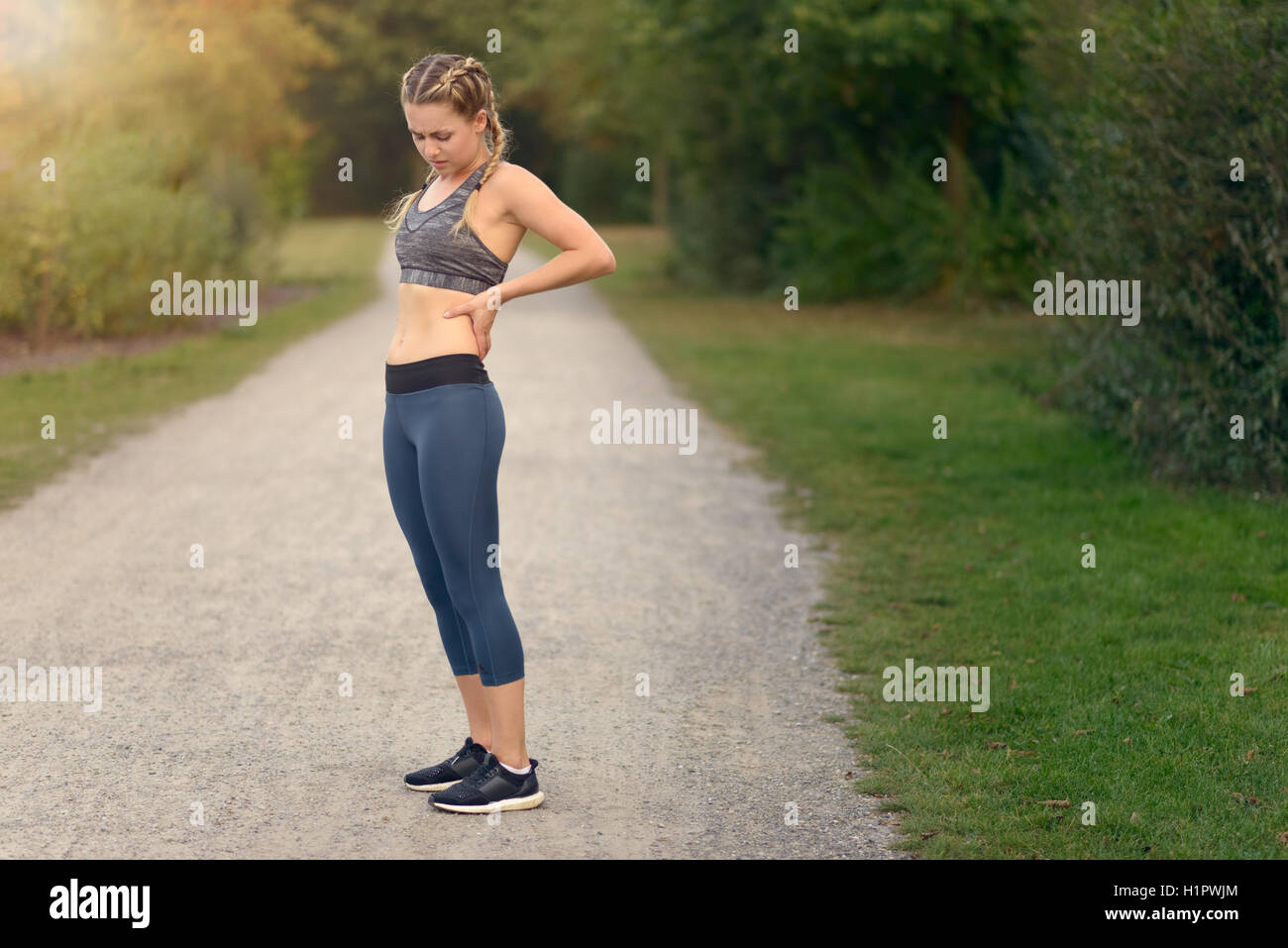 Woman athlete pausing to relieve her back pain holding her hand to her lower back with a grimace while out training in the count Stock Photo