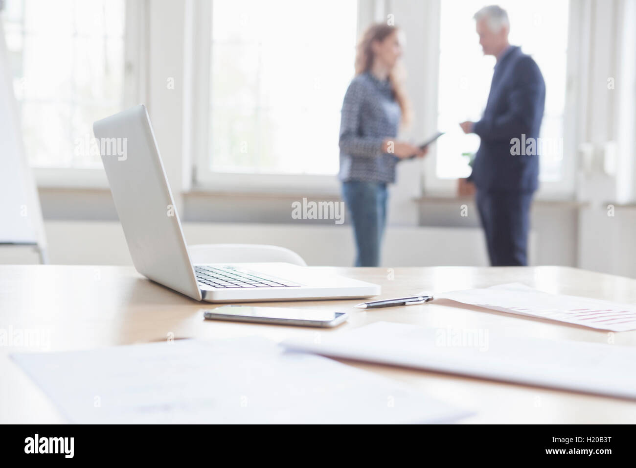 Laptop and documents on desk with businesspeople in background Stock Photo