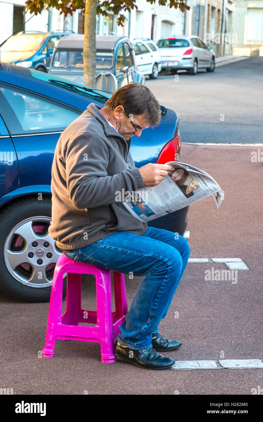 Man sitting on plastic stool reading newspaper - France. Stock Photo