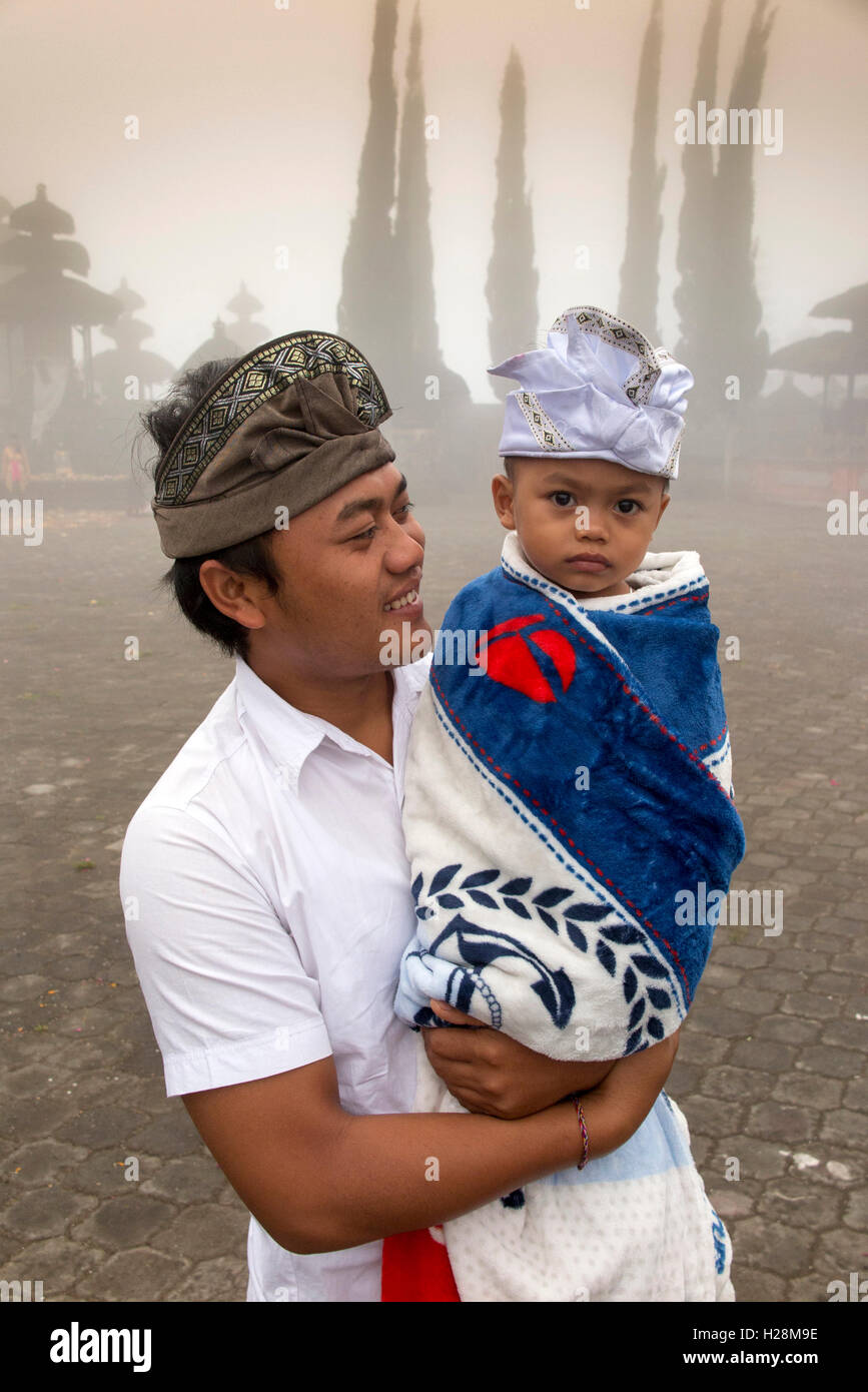Indonesia, Bali, Batur, Pura Ulun Danu Batur, man wearing traditional udeng hat carrying young son wrapped in blanket Stock Photo