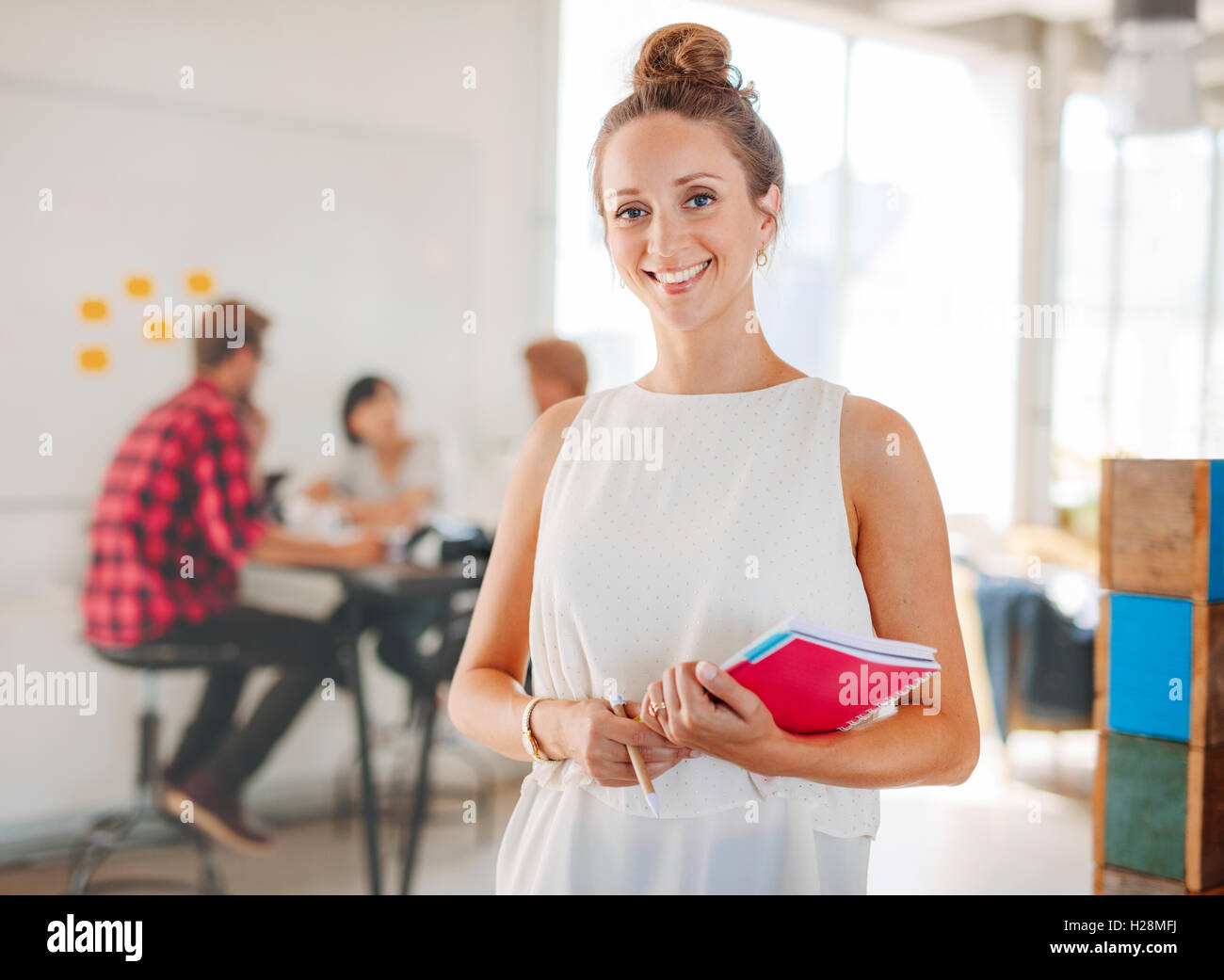 Portrait of beautiful young woman holding notepad with colleagues meeting in background. Business woman in casuals standing in m Stock Photo