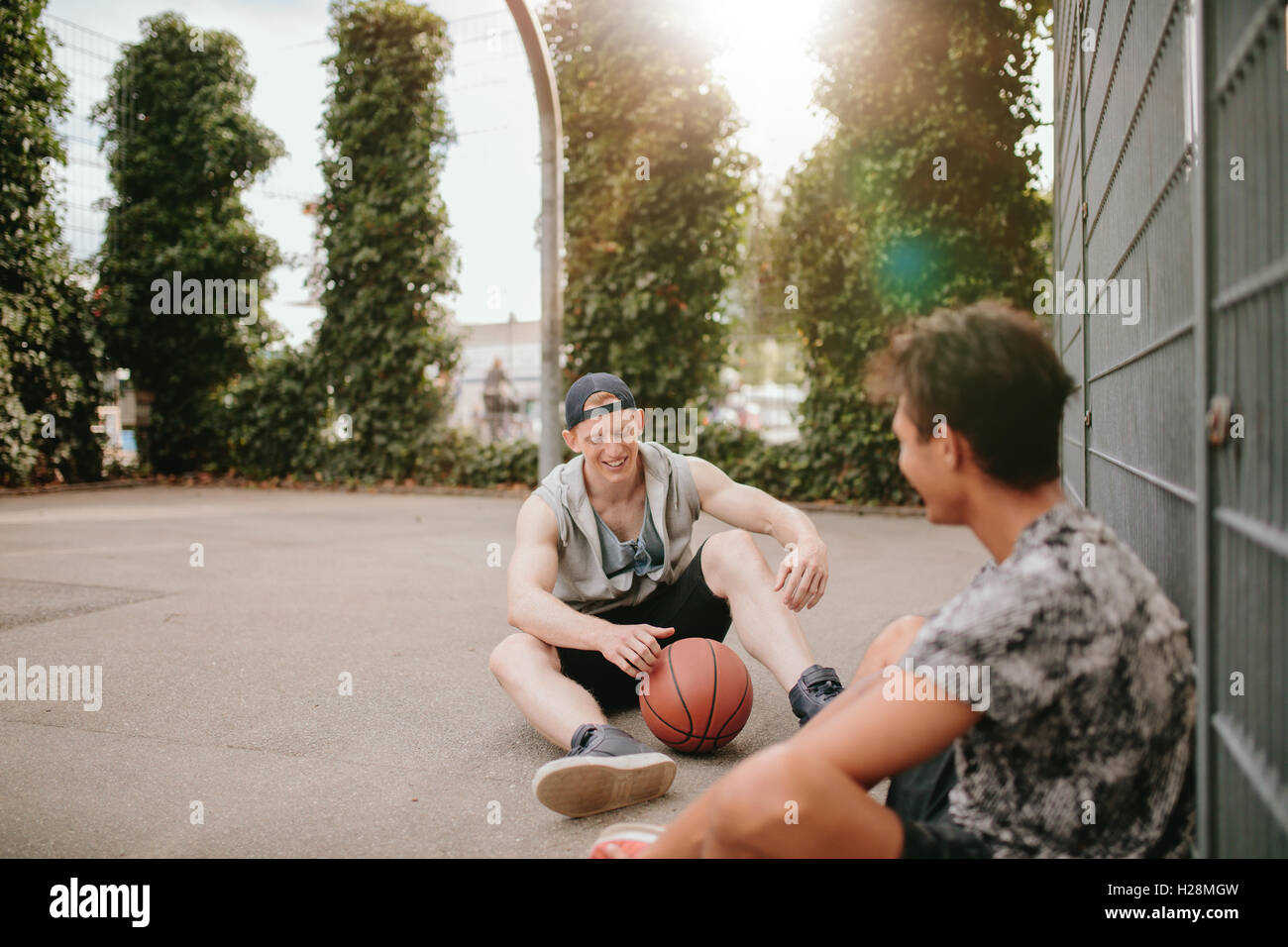 Two young men sitting on basketball court and smiling . Streetball players taking rest after playing a game. Relaxing and having Stock Photo