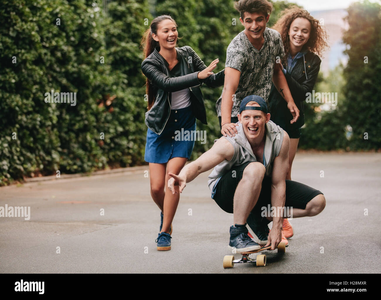 Full length shot of young men on skateboard with women friends pushing them. Group of teenagers enjoying outdoors with skateboar Stock Photo