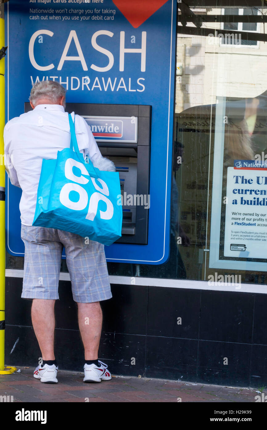 A senior man wearing shorts and trainers carrying a Co Op bag withdraws cash from a Nationwide Building Society ATM Cash Machine Stock Photo