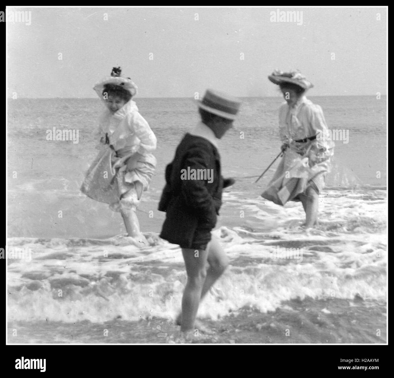 Two carefree women frolic in the waves of an English beach to the amusement of a young boy. Circa 1910. Photo by Tony Henshaw Stock Photo