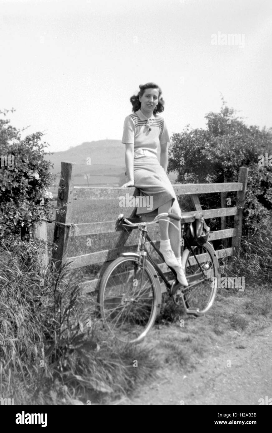 A young woman poses on a gate with her bicycle c1935. A lovely pose dressed in stylish knitted fashion with nice even lighting. Photo by Tony Henshaw Stock Photo
