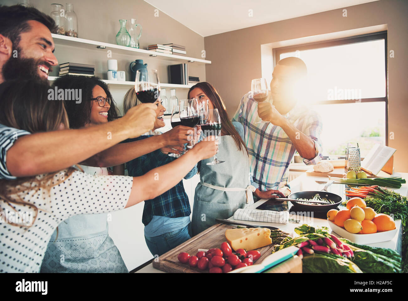 Group of people toasting wine glasses while preparing food for meal at large kitchen counter indoors Stock Photo