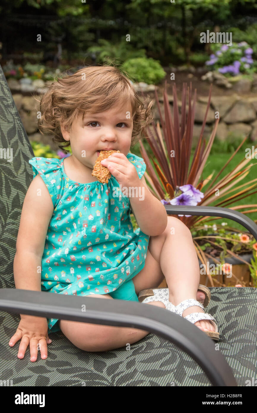 Eighteen month old girl sitting in a patio chair outside eating a granola bar in Issaquah, Washington, USA Stock Photo