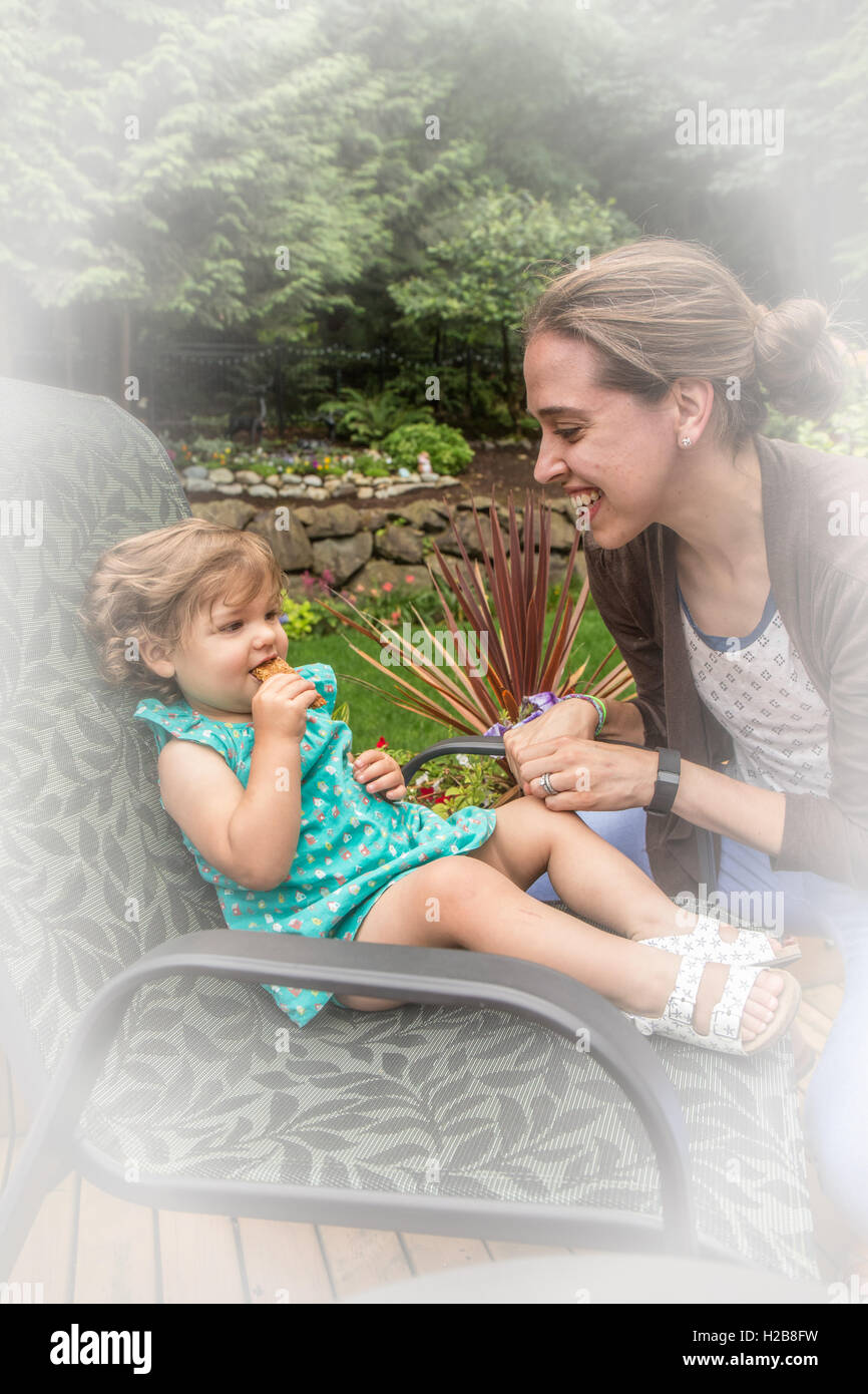 Eighteen month old girl eating a granola bar outside with her mother playfully teasing her, in Issaquah, Washington, USA Stock Photo
