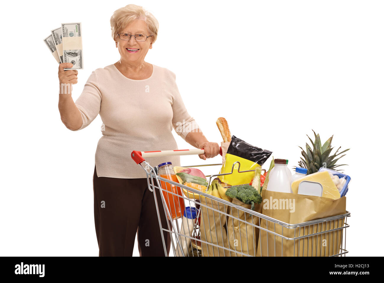 Happy elderly woman posing with a shopping cart full of groceries and stacks of money isolated on white background Stock Photo