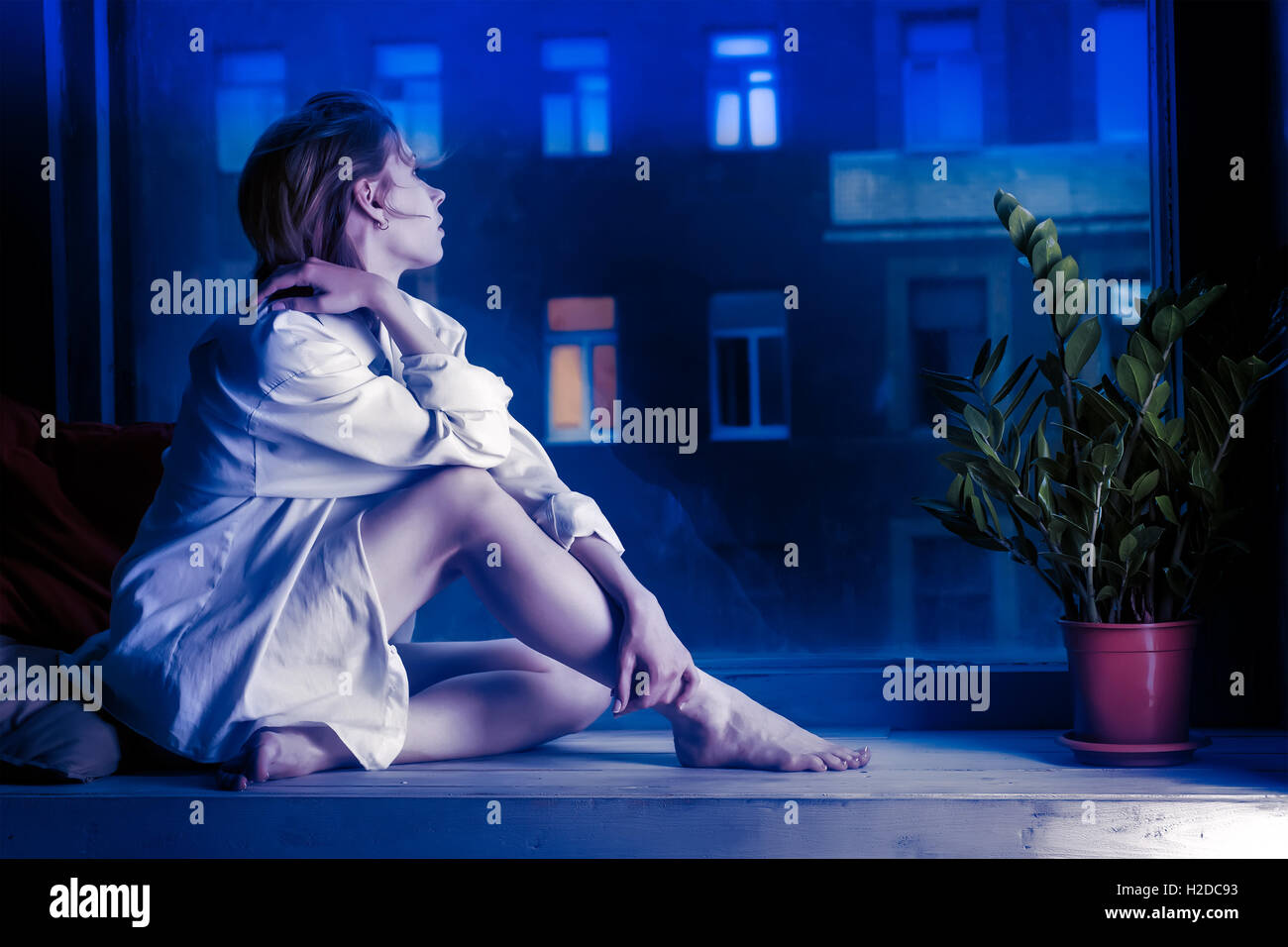 Half-dressed meditative girl in white shirt sits on window sill. Evening urban scene outside the window. Toned in blue colors Stock Photo