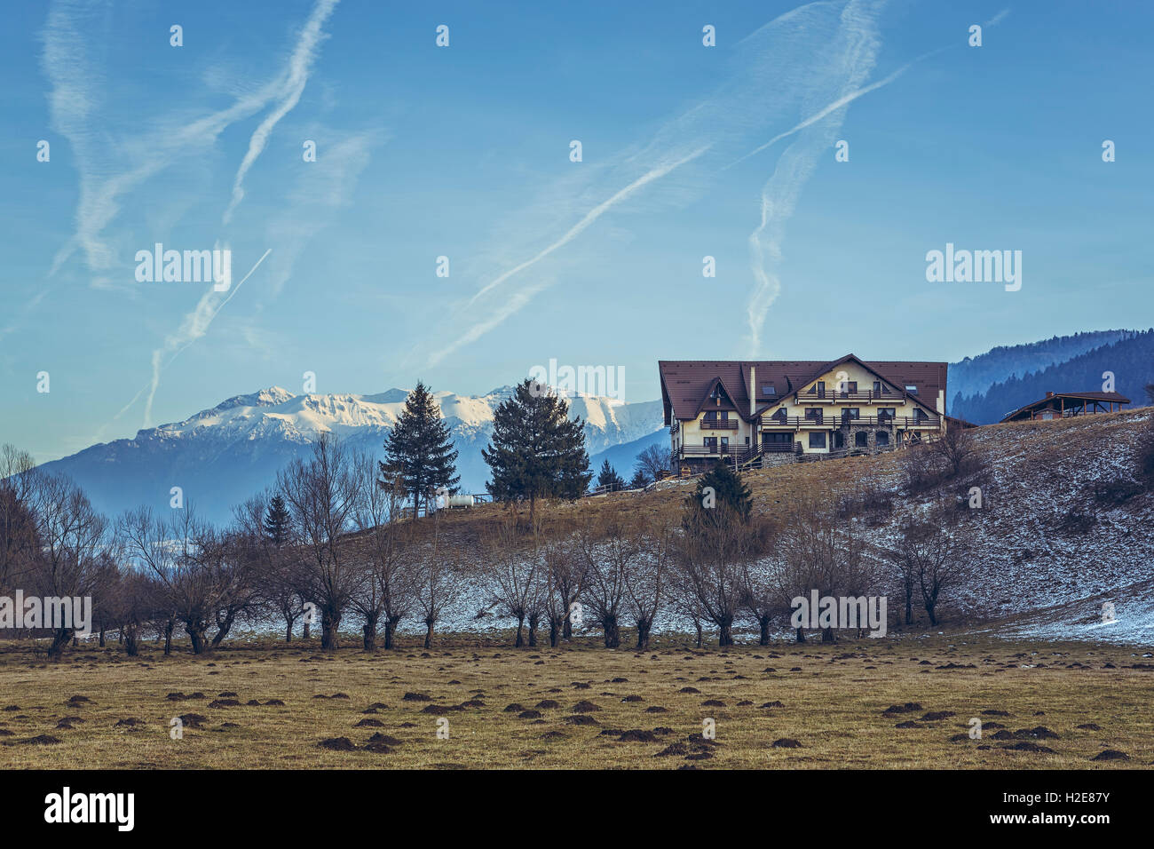 Scenic winter mountain landscape with remote modern pension, snow capped Bucegi mountains ridge at the horizon and lots of jet p Stock Photo