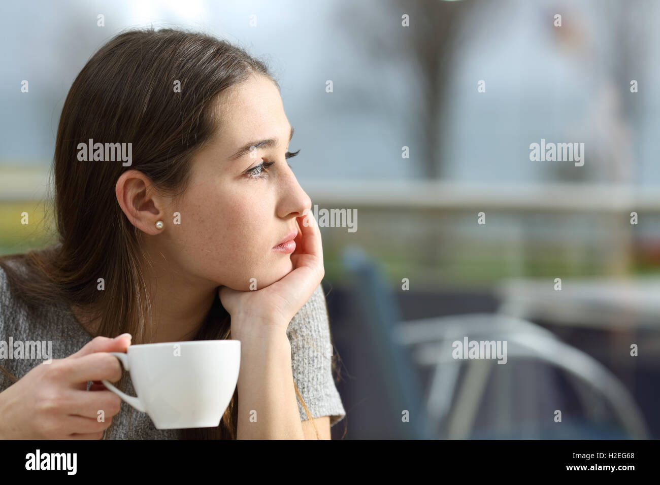 Pensive woman holding a coffee cup looking away in a restaurant terrace in a rainy day Stock Photo