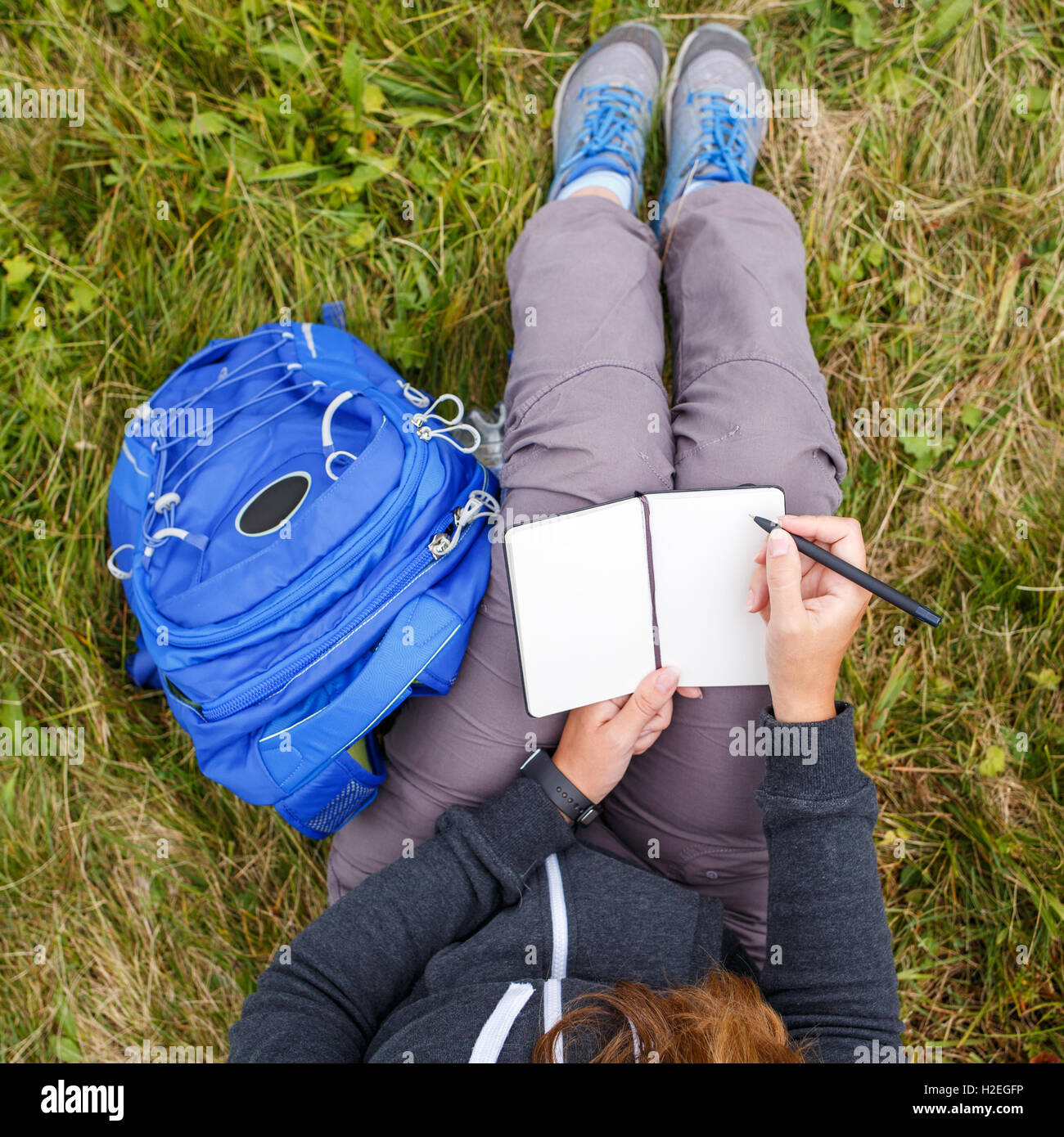 Woman with blue backpack sitting on grass and writing into blank notebook. Overhead view Stock Photo