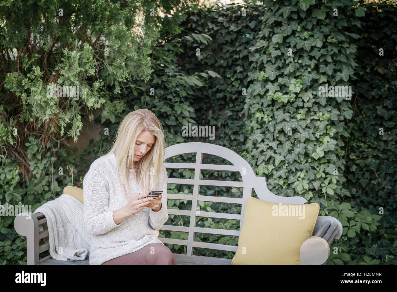 Woman sitting in a garden on  a bench, using her mobile phone. Stock Photo