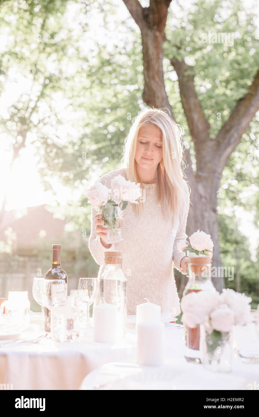 blonde woman setting a table in a garden, candles and vases with pink roses. Stock Photo