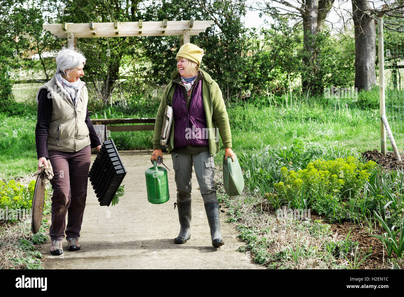 Two women walking along a path carrying kneeler pads and watering can and plant trays. Stock Photo