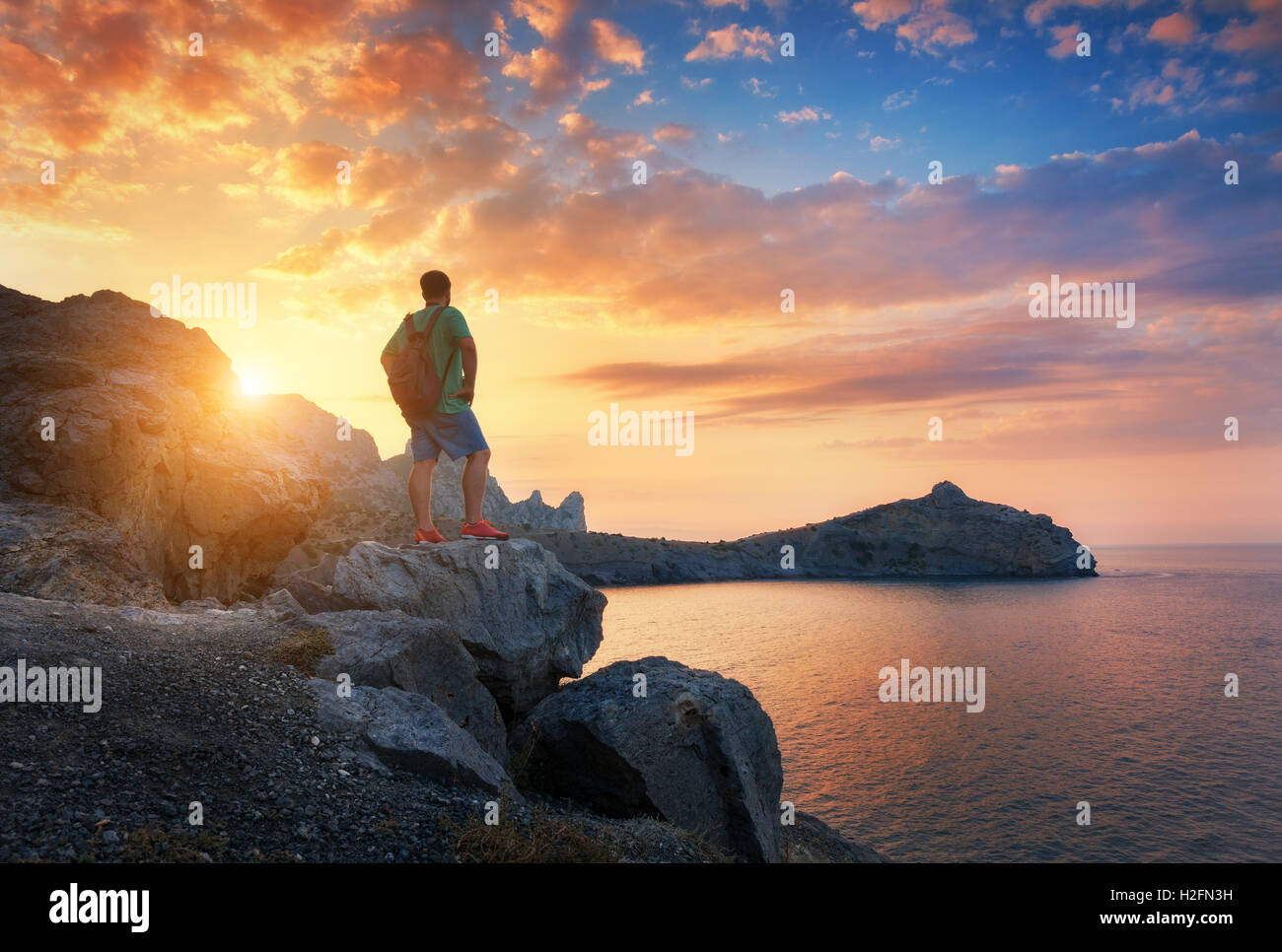 Beautiful summer landscape with standing man with backpack on the stone at the ocean against the colorful sky with clouds at sun Stock Photo