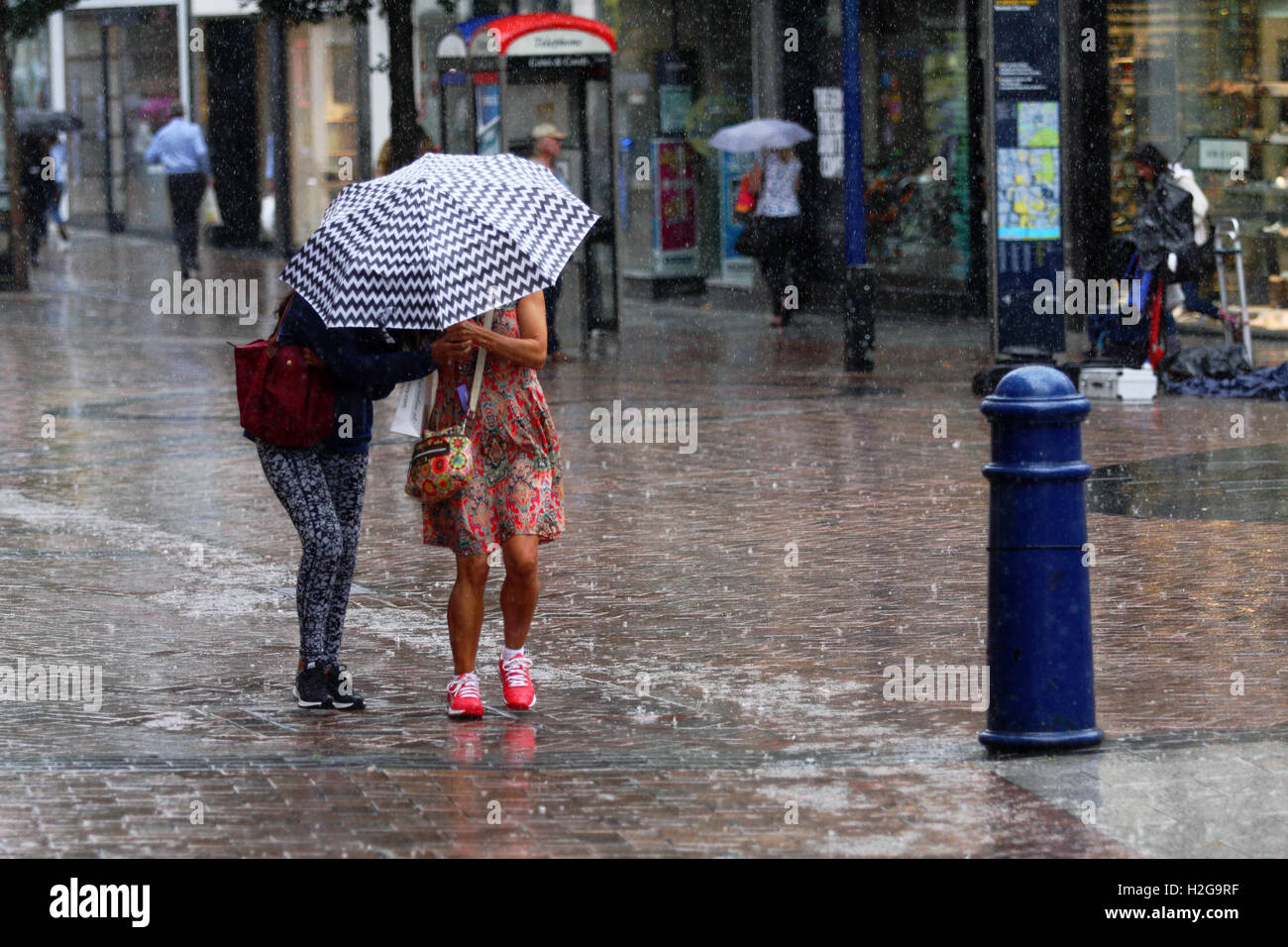 Two females caught in a sudden shower of rain try to stay dry under an umbrella in Kingston, Surrey, England. Stock Photo