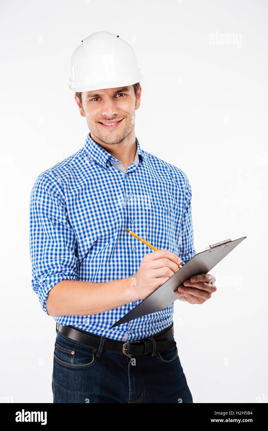 Smiling young man architect in building helmet standing and writing on clipboard Stock Photo