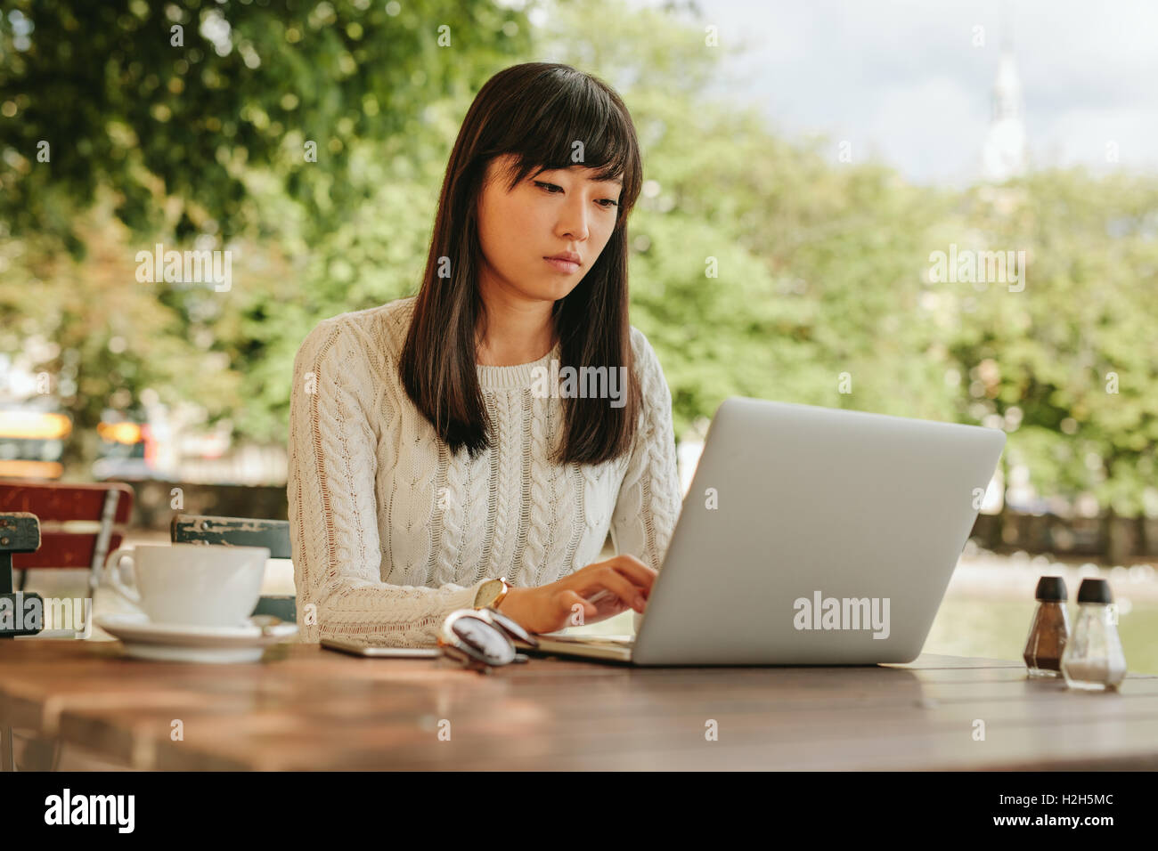 Shot of attractive young woman sitting at cafe table outdoors and working on laptop. Asian female surfing internet on laptop com Stock Photo