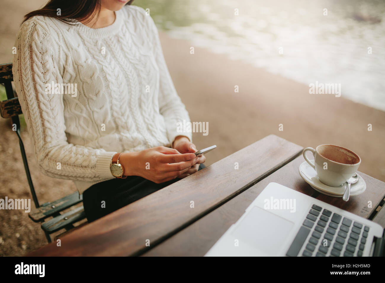 Cropped shot of young woman sitting at outdoor cafe using cellphone. Female sitting at cafe table with laptop and mobile phone i Stock Photo