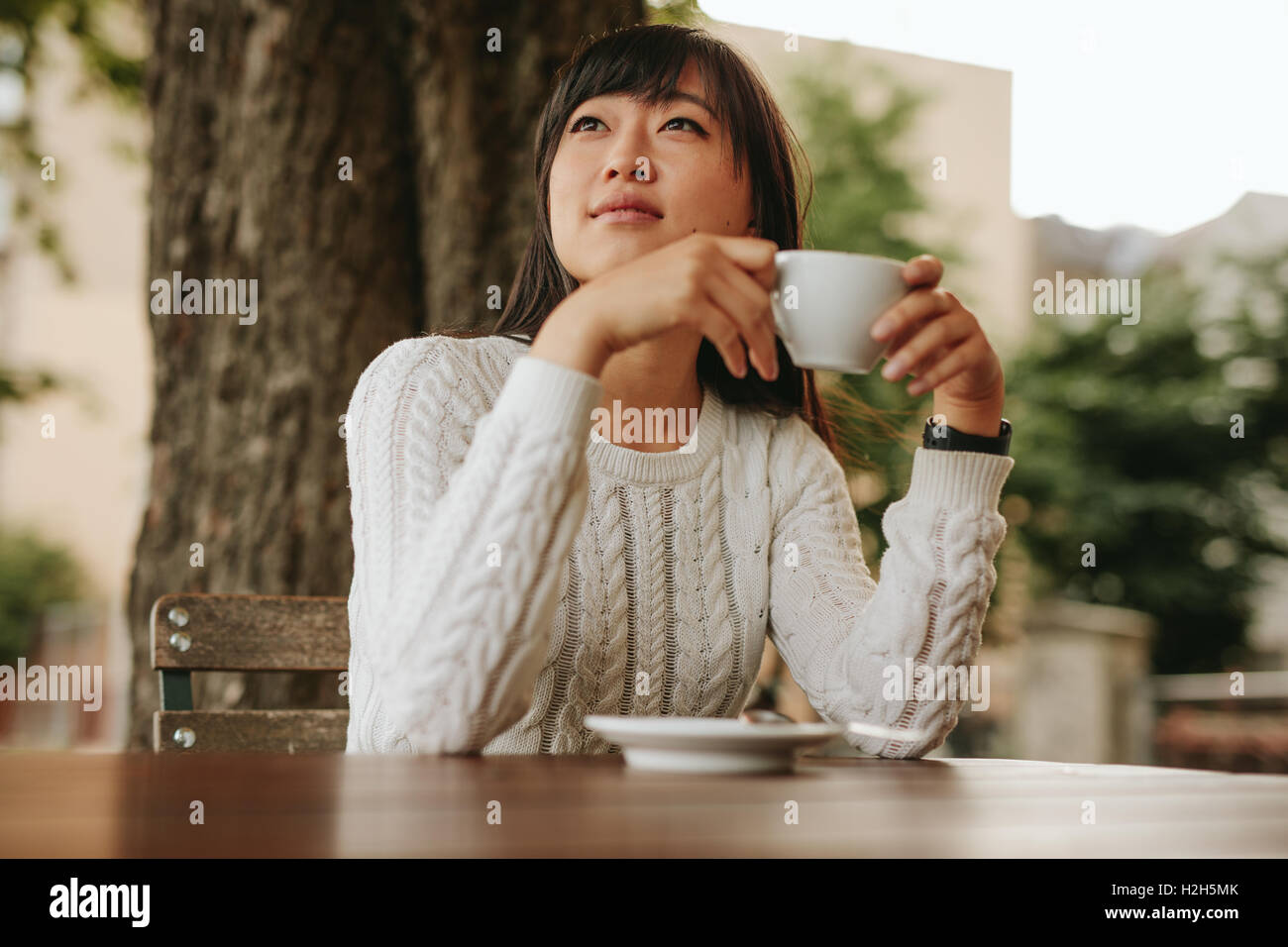 Young woman holding a cup of coffee and looking away. Chinese female drinking coffee at cafe. Stock Photo