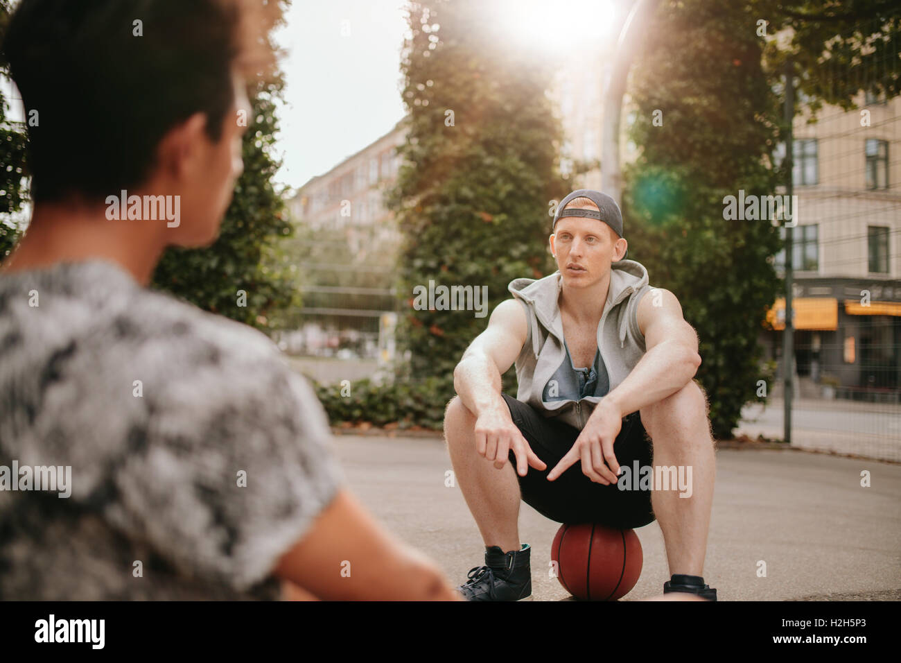 Young man sitting on top of basketball on court and talking with a friend. Streetball players taking break after playing a game. Stock Photo