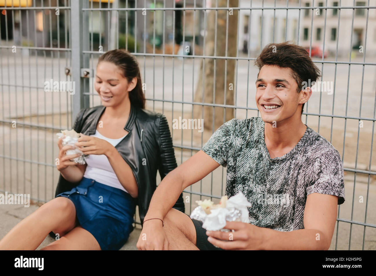 Young man and woman sitting outdoors and smiling. Teenagers relaxing outdoors and eating. Stock Photo