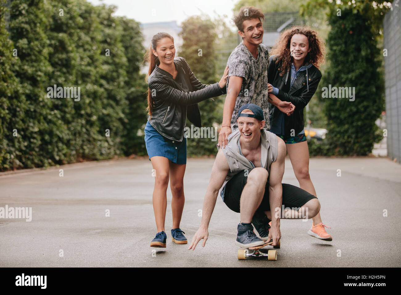 Full length shot of young men on skateboard with women friends pushing. Four teenagers enjoying outdoors with skateboard. Stock Photo